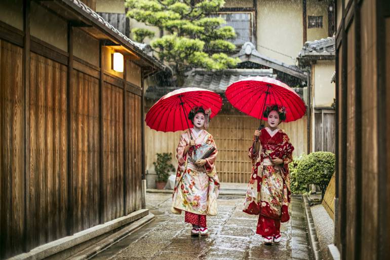 Two Maiko in ornate kimono holding red wagasa umbrellas walking a narrow Kyoto lane