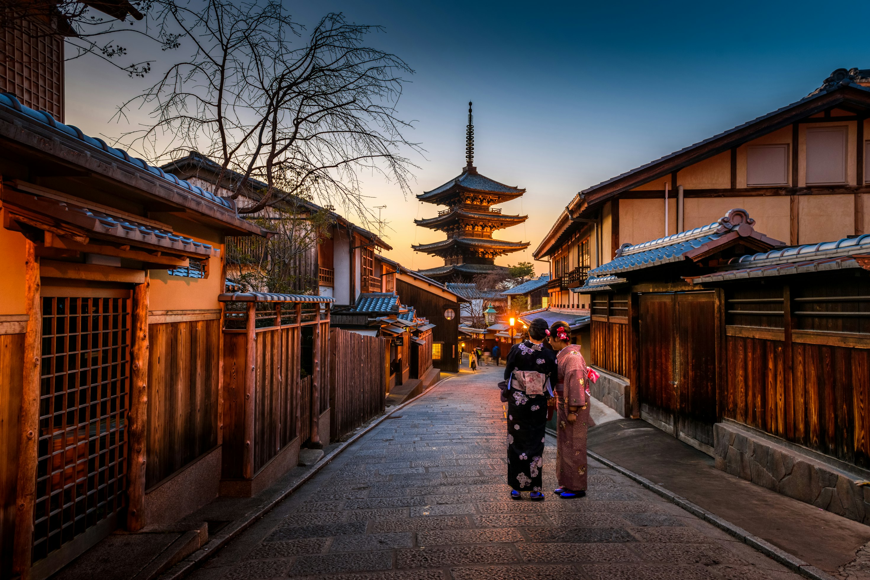 Two women in kimono walking Ninenzaka at dusk with Yasaka Pagoda lit ahead.