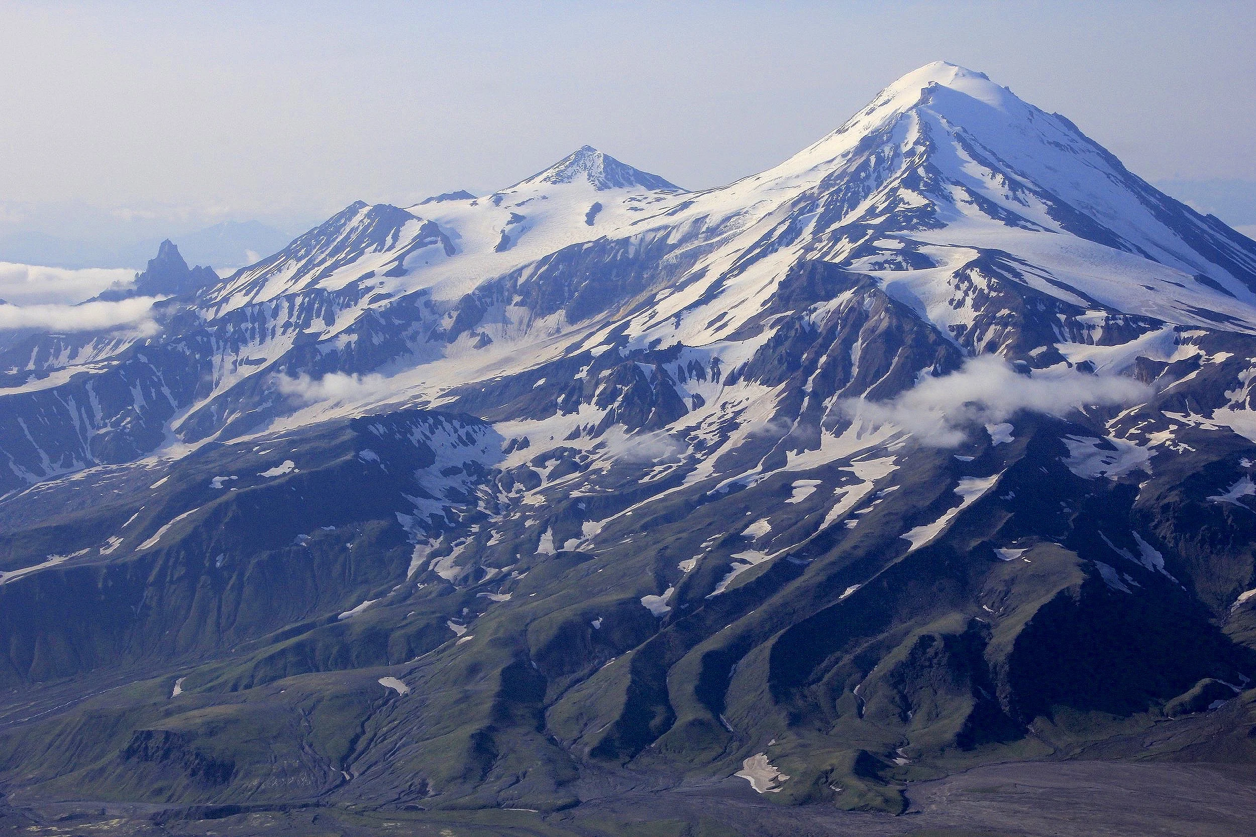 Snow‑clad stratovolcano with sharp summit and radial gullies above green foothills.