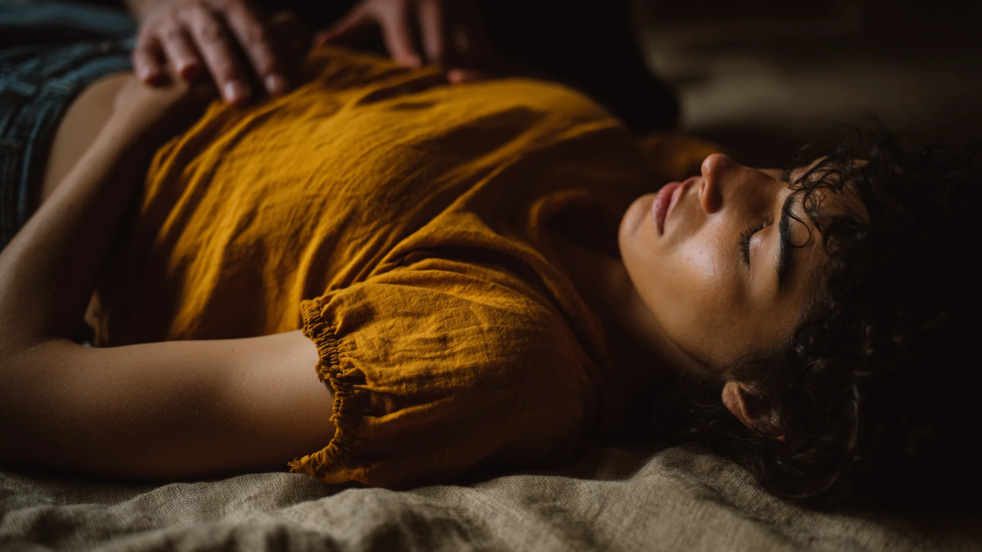 Person lying on floor, eyes closed, guide's hand on lower back ribs during gentle breath work.