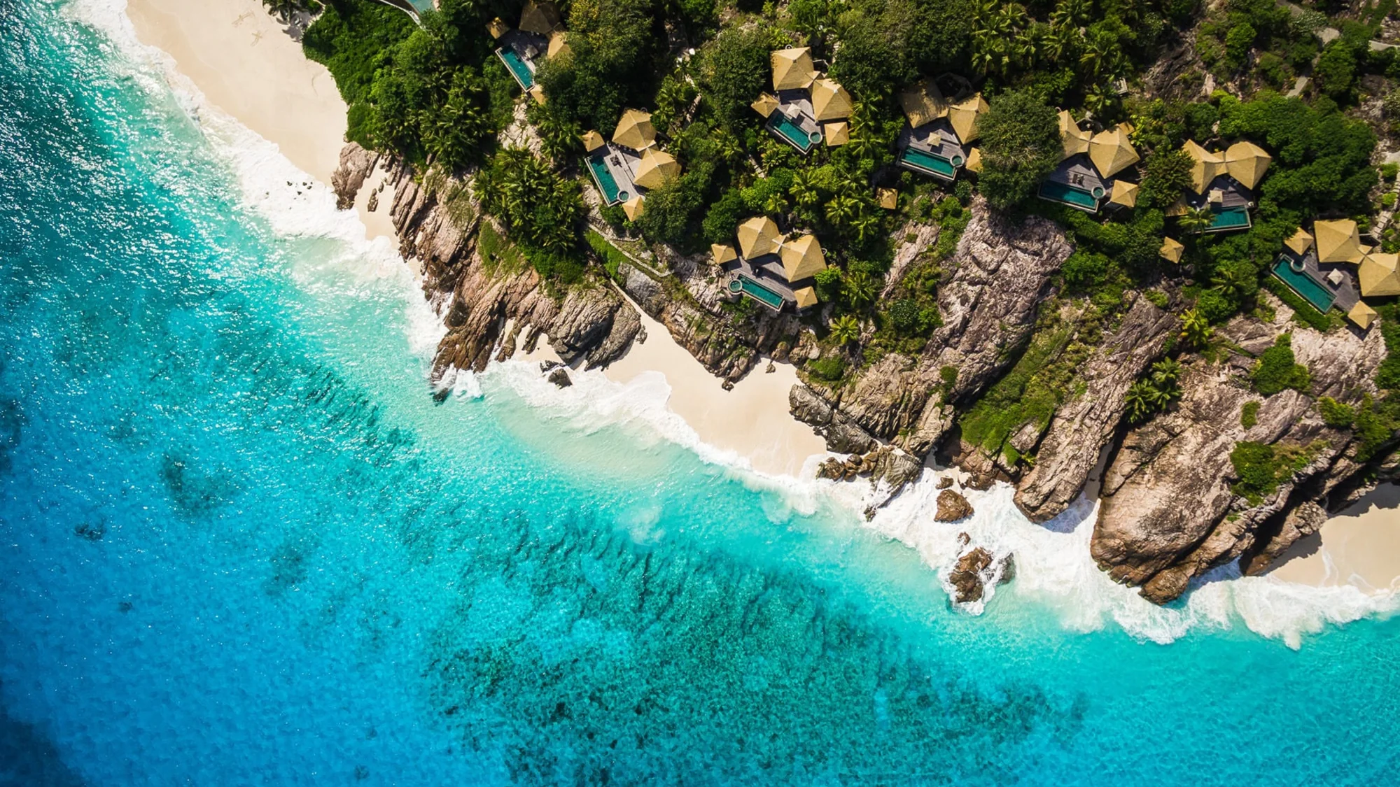An aerial view of a coastline with clear blue water, sandy beach, and tropical villas on rocky terrain.