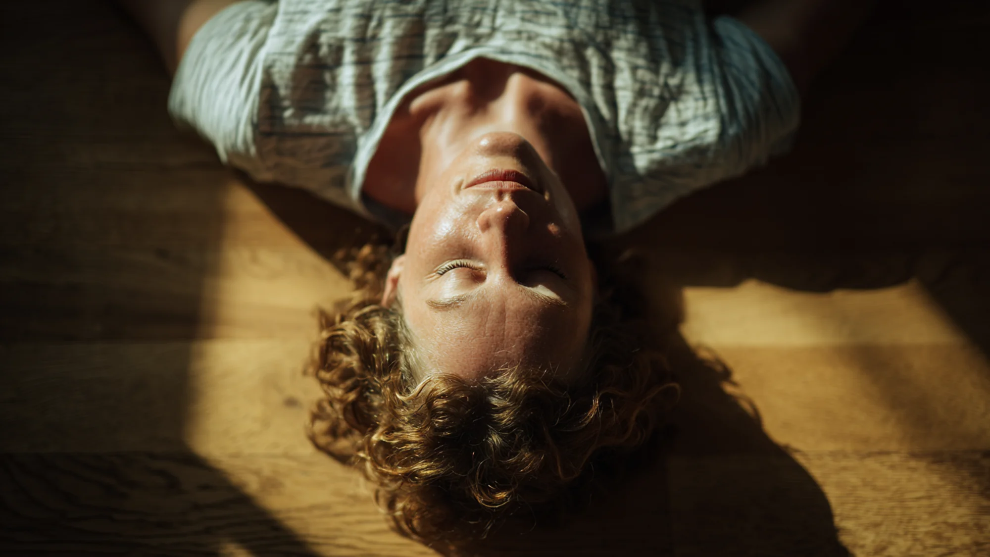 Person lying on back on wooden floor, arms relaxed, open receptive posture, natural overhead light.