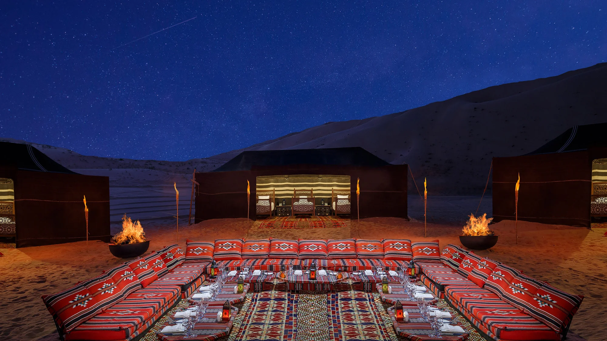 A desert dining setup with colorful cushions, lanterns, and tents under a starry sky.