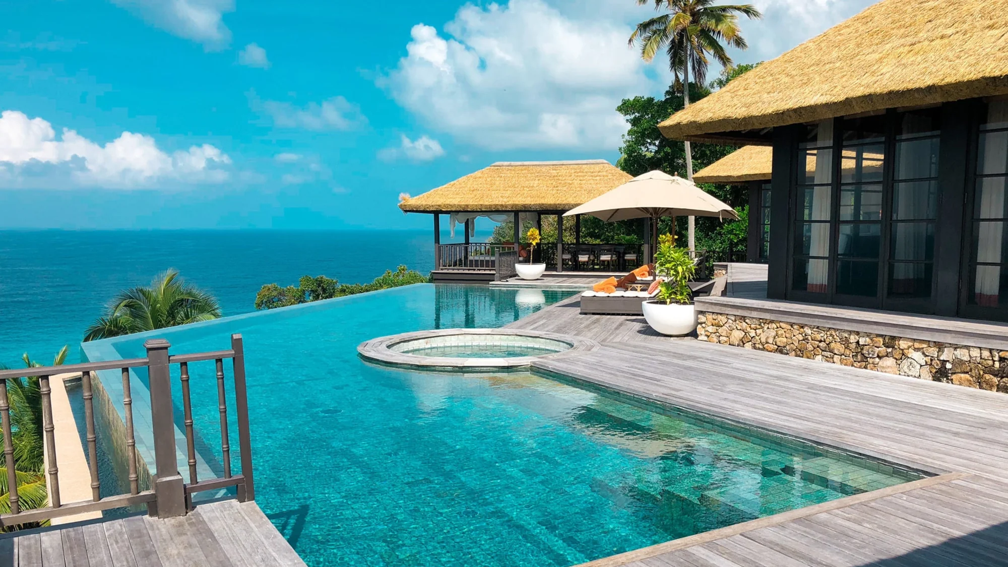 A luxurious villa with an infinity pool overlooking the ocean, surrounded by palm trees and a clear blue sky.
